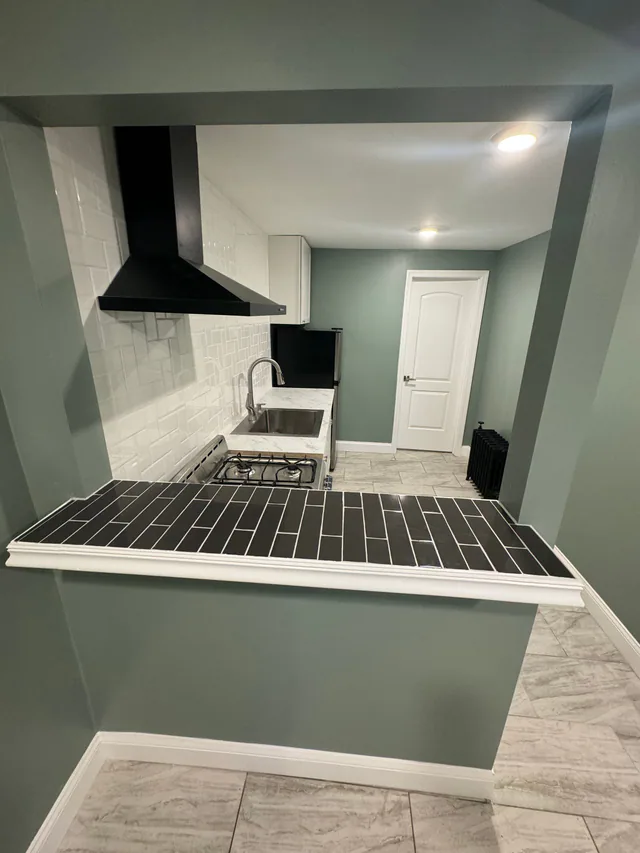 Finished kitchen viewed over a tile-topped half wall, with black hood, subway tile backsplash, gas range, and radiator